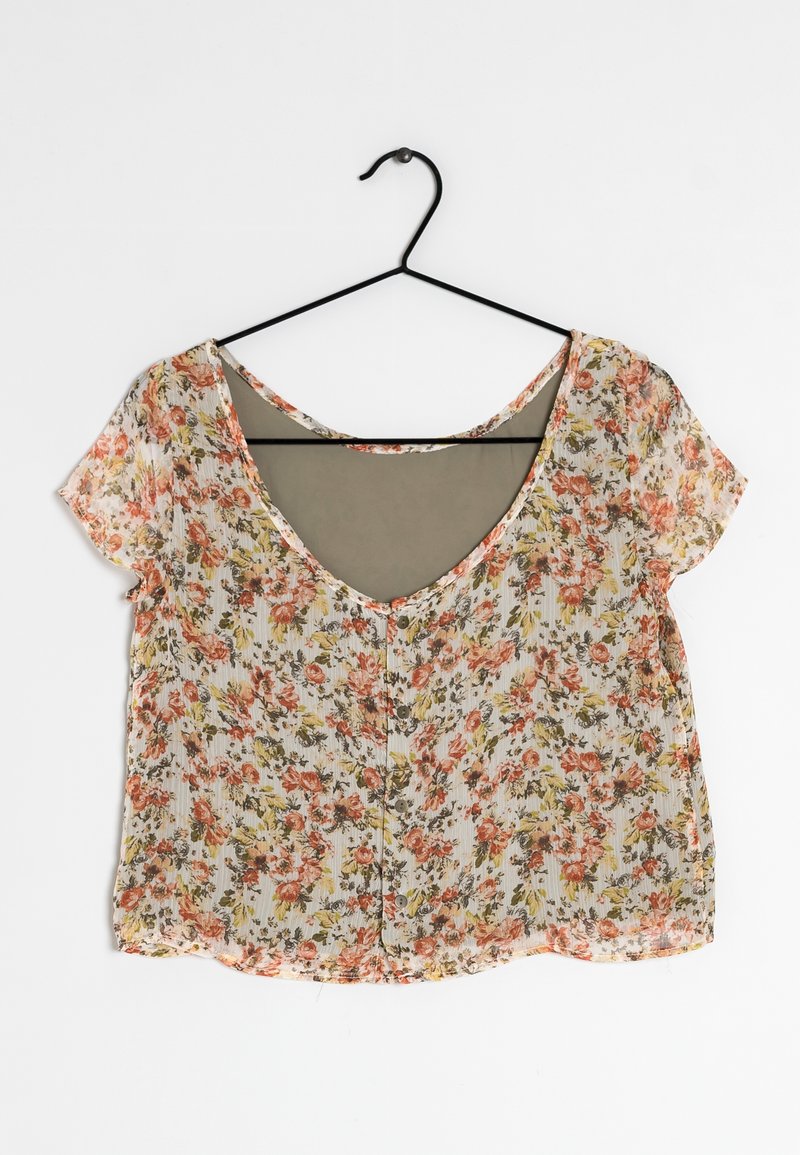 Blouse à manches courtes à motif floral avec des fleurs roses et jaunes, boutonnée devant, suspendue sur un cintre noir sur fond blanc.