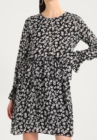 Mustapuku, joka on valmistettu kevyestä kankaasta, jossa on valkoisia ja harmaita kukkakuvioita. Pitkät hihat, röyhelövyötärö ja pyöreä pääntie.