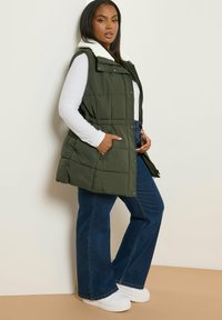 Zielona kamizelka z ociepleniem i białym futrzanym kołnierzem, z zapięciem na guziki, bocznymi kieszeniami i luźnym fasonem, noszona na niebieskich dzwonach i białych sneakersach.