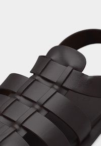 Sandale en cuir noir avec larges brides tissées et un design à bout arrondi sur un fond clair uni.