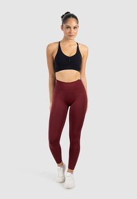 Musta urheilurintaliivi, jossa rypytetty etuosa, yhdistetty korkea vyötäröisiin, sileästä ja joustavasta kankaasta tehtyihin marjapuunpunaisiin leggingsseihin. Valkoiset urheilukengät.