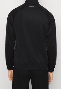 Chaqueta deportiva negra con un alto cuello, mangas raglán y sutiles acentos grises. Presenta una cinturilla y puños elásticos; con la marca "VENUM" en la parte posterior.