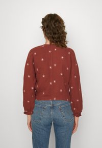 Rote strukturierte Bluse mit langen Ärmeln, verziert mit floraler Stickerei in helleren Farbtönen. Kombiniert mit blauen hochgeschnittenen Jeans.