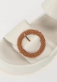 White faux leather sandal with a chunky sole, featuring a round, beaded brown buckle accent. Smooth surface with stitched edges.