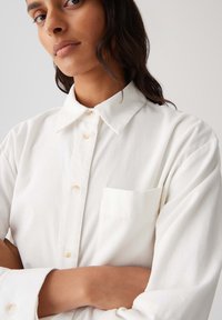 Vrouw met gekruiste armen, gekleed in een witte blouse met knopen en een voorzak, gefotografeerd tegen een effen lichte achtergrond.