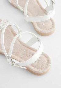 Sandali piatti bianchi con più fasce, caratterizzati da un accento glitter e una suola interna beige morbida. La suola esterna è marrone chiaro e testurizzata.