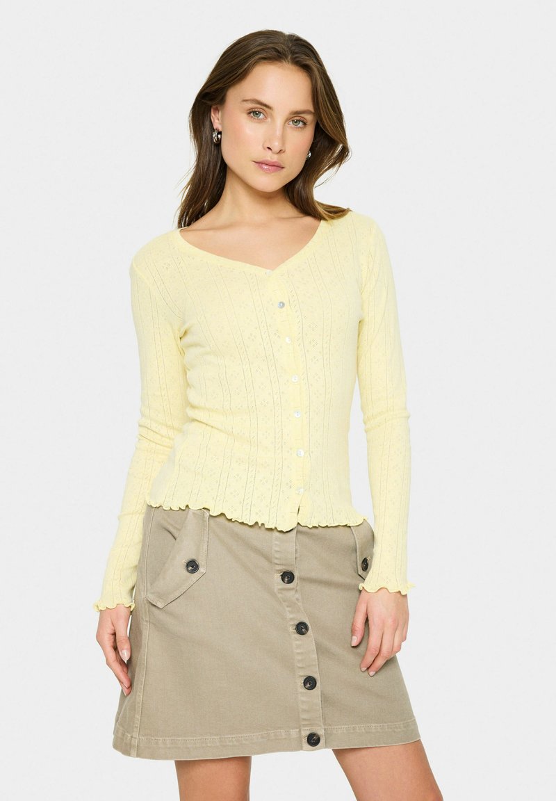Ljusgul stickad cardigans med V-hals, knäppning fram och subtilt mönster; tillsammans med en beige knäppskjorta i midjan med fickor.