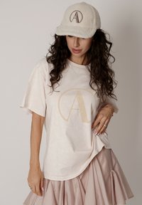Jasny t-shirt z krótkim rękawem z dużym beżowym logo "A", zestawiony z puszystą beżową czapką i jasnoróżową warstwową spódnicą.