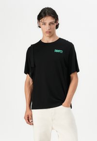 Camiseta negra de manga corta hecha de tela suave, con un logo verde "ROOTS" en el pecho izquierdo y un corte relajado.
