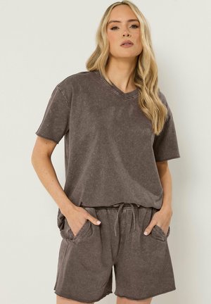 Femme aux longs cheveux blonds portant un t-shirt décontracté à col en V marron délavé et un short assorti à cordon avec poches, les mains dans les poches.