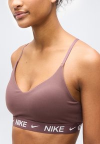Mladá osoba nosící fialovorůžovou sportovní podprsenku Nike s tenkými ramínky a elastickým páskem s logem pod prsy, na jednobarevném světlém pozadí.