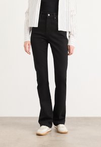 Jeans noirs à coupe slim, en denim. Comprend cinq poches et une fermeture à bouton sur le devant. Portés avec un haut noir et une chemise rayée.