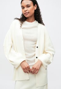 Γυναίκα με σγουρά μαλλιά που φοράει ένα υπερμεγέθες κρέμ κασκόλ πάνω από ένα ανοιχτό μπεζ πουλόβερ και αντίστοιχη ριμπ φούστα, απλωμένη σε μια μονόχρωμη φόντο.