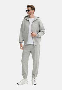 Felpa grigia con zip frontale e tasche, abbinata a pantaloni tuta coordinati. Indossata sopra una maglietta bianca. Scarpe sportive bianche, occhiali da sole indossati.