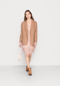 Blazer castanho sobre um vestido floral com babados, combinado com botins castanhos e uma mala creme. Tecido suave, design elegante, cores suaves.