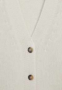 Cardigan tricoté couleur crème avec deux boutons marron et coutures visibles texturées.