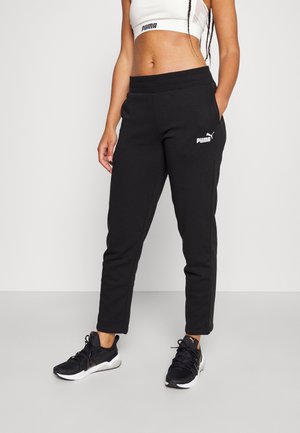 Femme portant un pantalon de survêtement Puma noir, des chaussures de sport noires et un crop top blanc Puma, debout devant un fond uni.