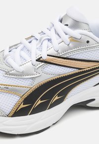 Zapatillas deportivas con parte superior de malla blanca, acentos en plata y oro, detalles en negro y cordones blancos. Superficie texturizada y collar acolchado.