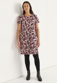 Kortärmad klänning i vinröd med ett abstrakt beige bladmönster, som har sidofickor och en avslappnad passform. Tillsammans med svarta leggings och ankelstövlar.