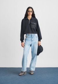 Μαύρο cropped φούτερ με φερμουάρ στον γιακά και τσέπη, ανοιχτό μπλε τζιν σε ίσια γραμμή, γκρι αθλητικά παπούτσια και σκούρο καπέλο. Το μοντέλο στέκεται πάνω σε μπλε χαλί.