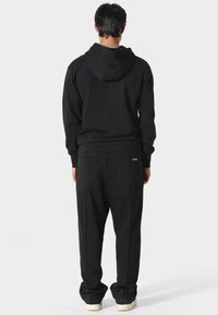 Hombre de pie mirando hacia atrás, vestido con una sudadera negra con capucha, pantalones negros con bolsillos traseros y zapatillas blancas, sobre un fondo claro y liso.