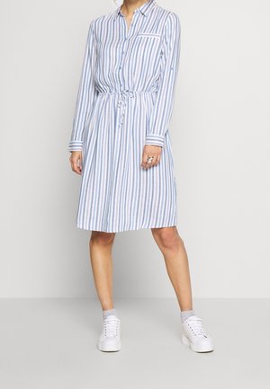 Kobieta ubrana w koszulową sukienkę o długości do kolan w pionowe niebiesko-białe paski, z długimi rękawami, w talii ze sznurkiem, białe trampki i szare skarpetki do kostek.
