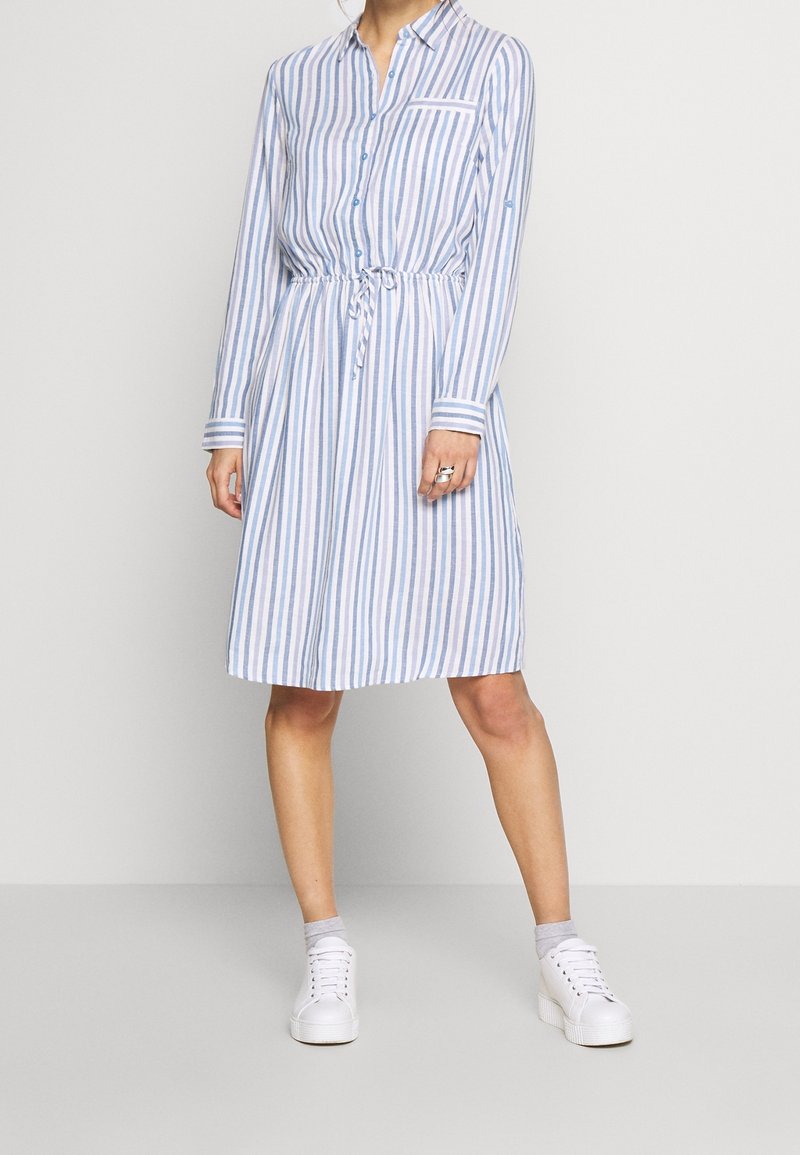 Femme portant une robe chemise à rayures verticales bleues et blanches, longueur au genou, manches longues, taille ajustée par un cordon, baskets blanches et chaussettes grises aux chevilles.