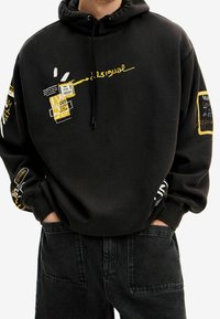 Felpa nera con accenti grafici gialli che presentano testo e motivi. Cappuccio con coulisse regolabile e polsini a coste. Indossata con jeans scuri.