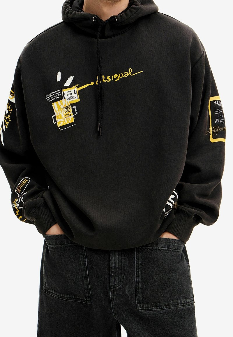Felpa nera con accenti grafici gialli che presentano testo e motivi. Cappuccio con coulisse regolabile e polsini a coste. Indossata con jeans scuri.