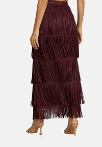 Mujer vestida con una falda de franjas escalonadas burdeos y sandalias de tacón beige, vista desde atrás con las manos relajadas a los lados.