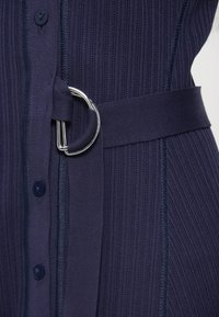 Tmavomodré pletené šaty zvýrazňujúce rebrovanú textúru, s tmavými gombíkmi, opaskom v páse so strieborným D-krúžkom a štruktúrovaným strihom.