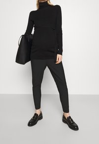 Person, der einen schwarzen Langarm-Turtleneck-Pullover und gestreifte Hosen trägt, eine große schwarze Handtasche hält und auf einem grauen Boden vor einer weißen Wand steht.