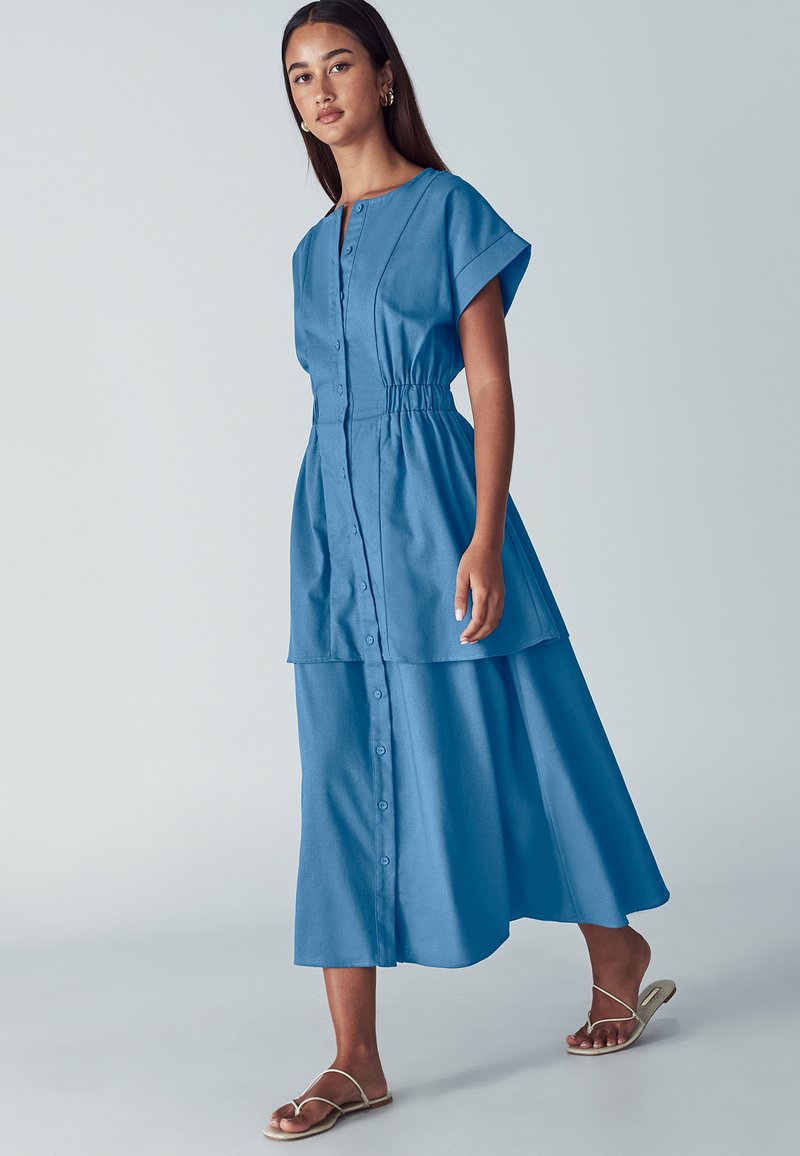 Svetlo modra, s kratkimi rokavi in gumbnim zapenjanjem, obleka z elastičnim pasom, ki vsebuje slojevito krilo in gladko teksturo tkanine.