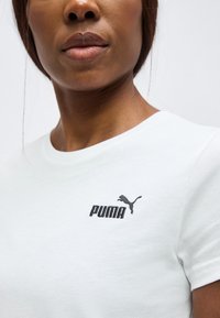 Gros plan d'une personne portant un t-shirt blanc Puma montrant le logo Puma sur la partie supérieure de la poitrine sur un fond uni.