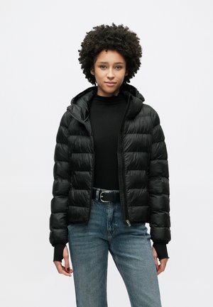 Doudoune noire avec fermeture éclair à l'avant, capuche et design matelassé. Le modèle porte un haut noir et un jean bleu, mettant en valeur la texture et la coupe de la veste.