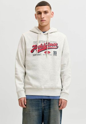 Młody mężczyzna w jasnoszarej bluzie Jack & Jones z czerwonym i czarnym logo oraz w niebieskich dżinsach na jednolitym, jasnym tle.