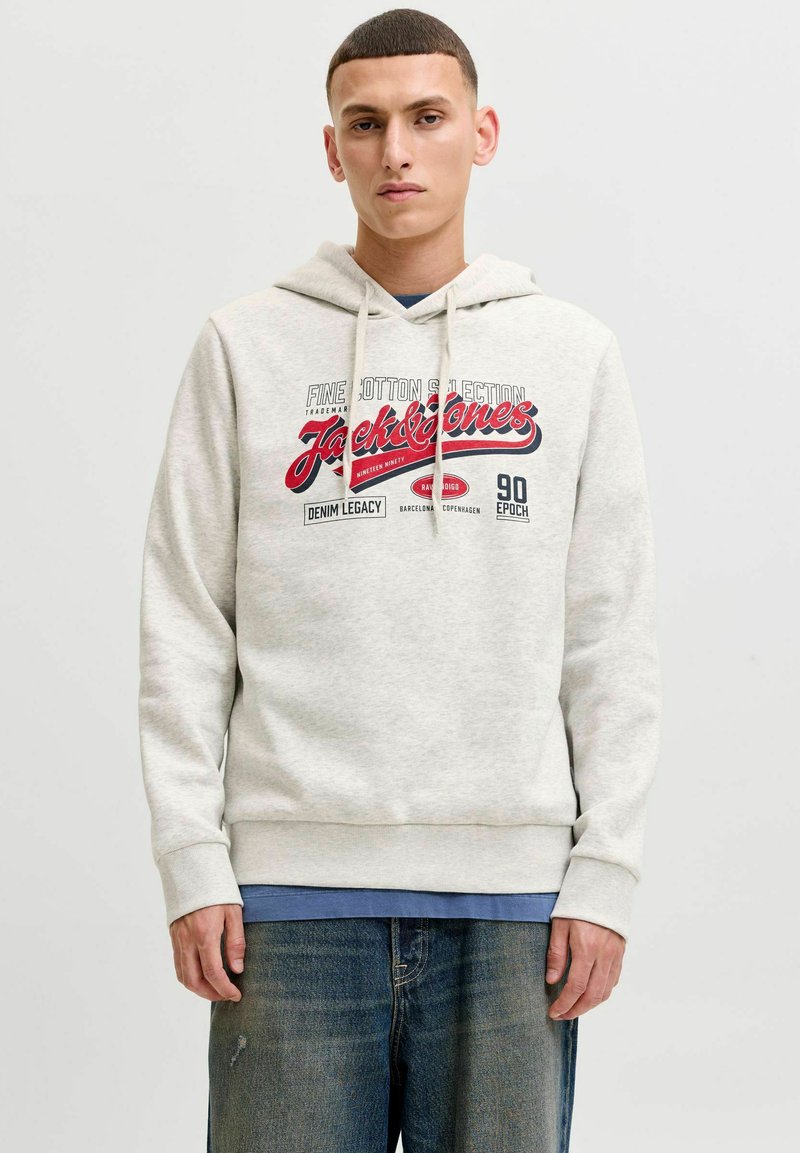 Νέος άνδρας φοράει ανοιχτό γκρι φούτερ Jack & Jones με κόκκινο και μαύρο λογότυπο και μπλε τζιν μπροστά σε ανοιχτόχρωμο φόντο.