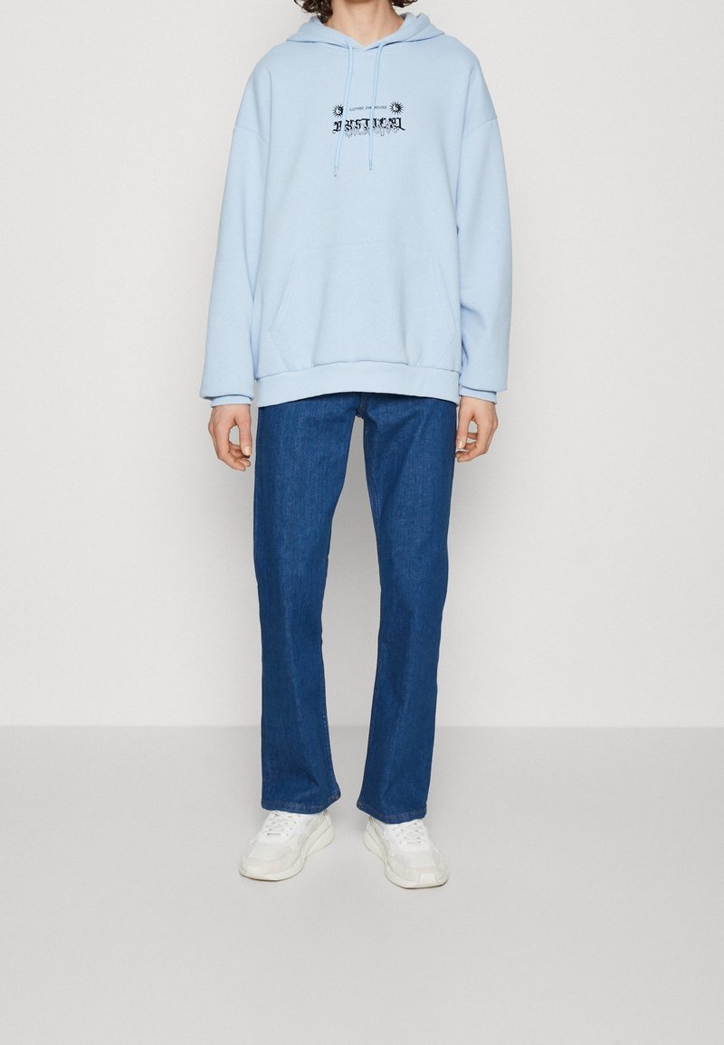 Jasnoniebieska bluza z kapturem z przednią kieszenią i czarną grafiką. Połączona z ciemnoniebieskimi jeansami o prostym kroju i białymi sneakersami. Miękka tkanina, luźny krój.