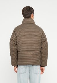Bruine en tan geruite pufferjack met hoge kraag, oversized pasvorm en elastische manchetten, gedragen over lichtblauwe spijkerbroek.