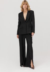 Czarny, dopasowany garnitur z marynarką o głębokim dekolcie w serek i szerokimi spodniami z przednimi rozcięciami. Uzupełniony o szpilki w cętki lamparta i złotą biżuterię.