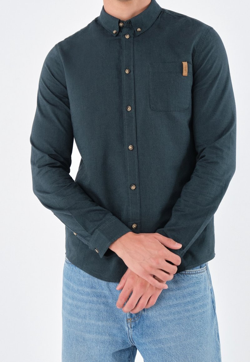 Homme portant une chemise boutonnée vert foncé avec une poche poitrine et un jean bleu clair, les mains croisées devant lui.