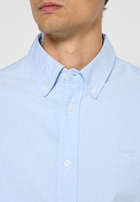 Camicia in cotone azzurro chiaro con colletto button-down, caratterizzata da bottoni bianchi e una trama sottile. Logo ricamato sul lato sinistro del petto.