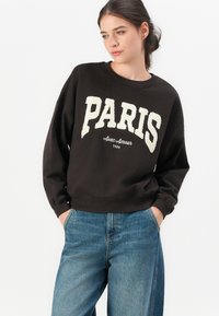 Woman with dark hair in a bun wearing a black sweatshirt with "PARIS Avec Amour 1986" text and blue jeans, hands in pockets, looking sideways.