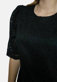 Čierny čipkovaný top s krátkymi pufovými rukávmi, s kvetinovou textúrou a okrúhlym výstrihom. Materiál vyzerá ľahko a jemne.
