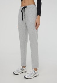 Pantaloni sportivi grigi chiari con cintura regolabile, design a gamba dritta e texture sottile. Indossati con scarpe da corsa argentate.