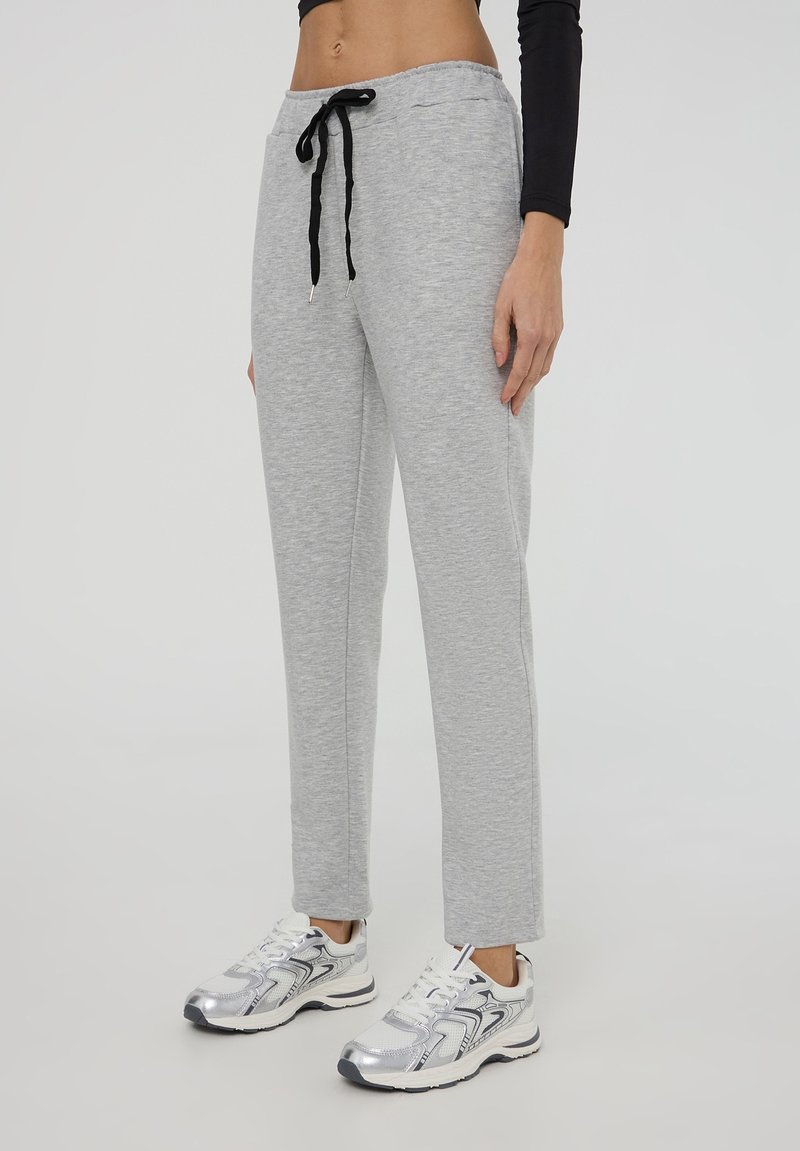 Pantaloni sportivi grigi chiari con cintura regolabile, design a gamba dritta e texture sottile. Indossati con scarpe da corsa argentate.