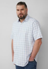Mężczyzna z brodą i związkami włosów, ubrany w jasnoniebieską kratkowaną koszulę z krótkim rękawem oraz dżinsy, stojący z rękami w kieszeniach, oparty o gładką ścianę.