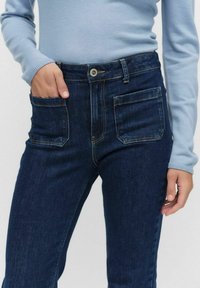 Tummansiniset korkeavyötäröiset farkut, joissa on kaksi etutaskua ja hillitty haalistuminen. Yhdistetty vaaleansiniseen pitkähiharaiseen puseroon, jossa on ribbikuvio.