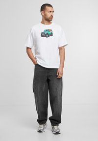 Hvid bomuld t-shirt med et farverigt grafisk motiv af en grøn lastbil med en hund indeni, parret med løstsiddende mørkegrå jeans og sneakers.