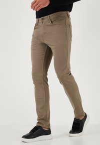 Kaki uske traperice slim fit s glatkom teksturom, s prednjim džepovima i suptilnim detaljem šivanja, uparene s crnim cipelama na vezivanje.